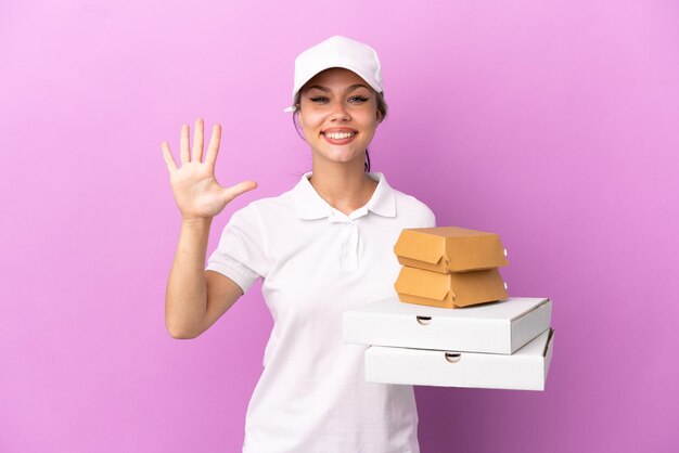 Consegna pizza Ragazza russa con uniforme da lavoro che raccoglie scatole di pizza e hamburger isolati su sfondo viola contando cinque con le dita