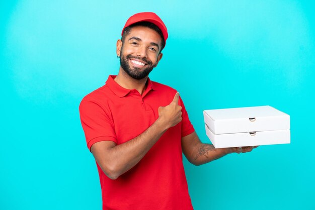 Consegna pizza Uomo brasiliano con uniforme da lavoro che raccoglie scatole di pizza isolate su sfondo blu che punta indietro