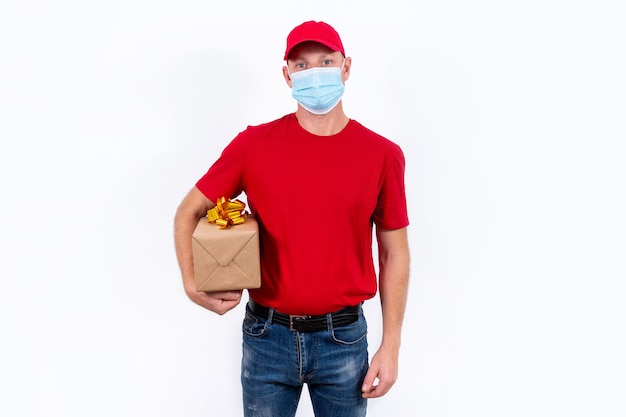 Consegna sicura di regali per le vacanze. Un corriere in uniforme rossa e maschera medica protettiva tiene la scatola con un fiocco. Ordini di regali a distanza senza contatto in quarantena durante la pandemia di coronavirus.