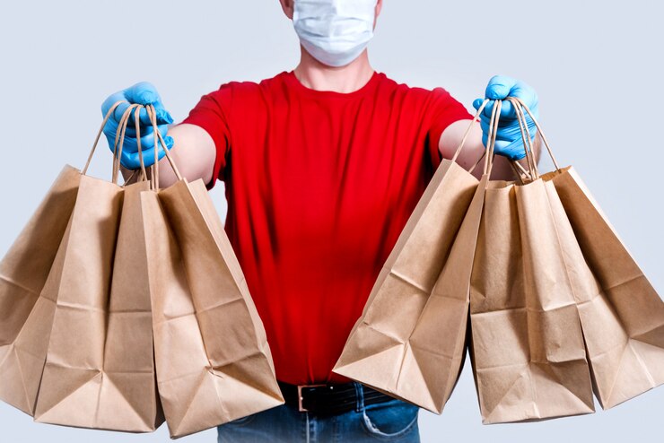 Consegna sicura. il corriere in uniforme rossa e maschera protettiva e guanti tiene un grosso ordine, molti sacchetti di carta, cibo per consegne senza contatto in quarantena.