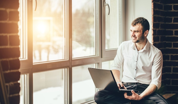 Considerato giovane uomo d'affari in camicia. Seduto con un laptop vicino alla finestra. Uomo che lavora con i documenti. Concetto di libero professionista. Resta a casa durante la quarantena. Pandemia.