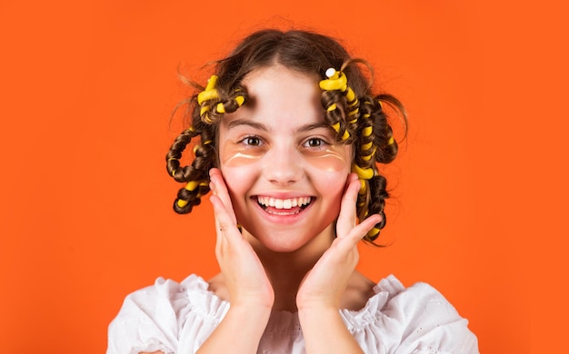 Consigli per lo styling Adolescenti hobby Piccola ragazza Arricciare i capelli Usare i bigodini Sfondo arancione Figlia con i bigodini in testa che ride Salone di parrucchiere Routine di bellezza femminile Acconciatura adorabile del bambino