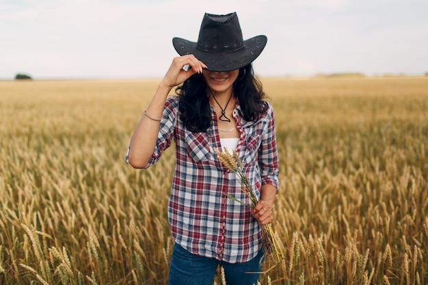 Contadina americana con spighe di grano covone che indossa cappello da cowboy, camicia a quadri e jeans al campo di grano.