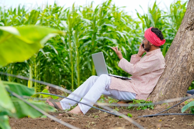 Contadino indiano che utilizza laptop e parla su smartphone al campo agricolo.