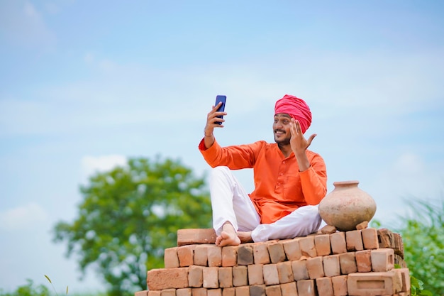 Contadino indiano seduto sui mattoni e utilizza lo smartphone al campo agricolo.