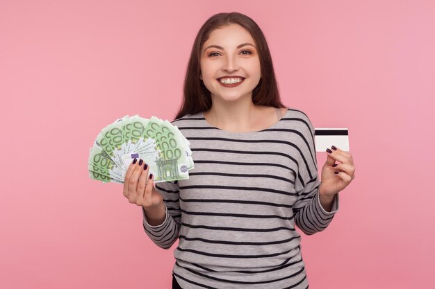 Contanti e denaro elettronico Ritratto di donna felice in felpa a righe con banconote in euro e carta di credito guardando la fotocamera con sorriso a trentadue denti soddisfatto girato in studio isolato su sfondo rosa