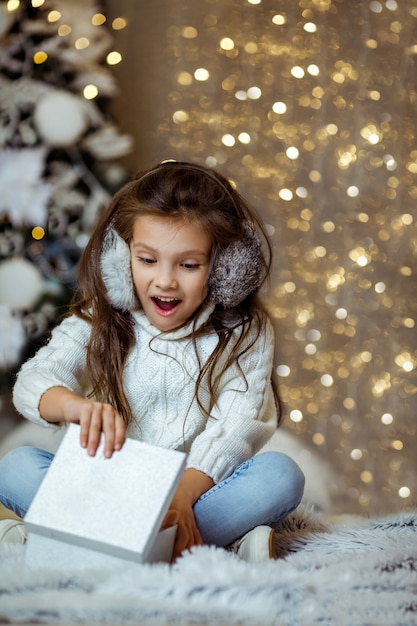 Contenitore di regalo di apertura della ragazza del bambino sorpreso felice vicino all'albero di Natale decorato con luci