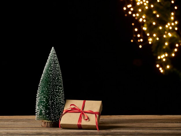 Contenitore di regalo di Natale e albero di abete sulla tavola di legno contro luci festive sfocate, spazio per testo