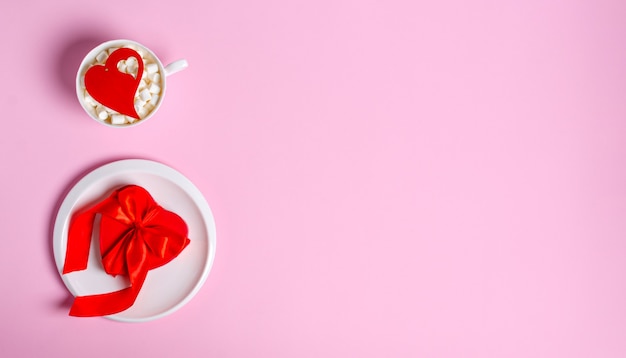 Contenitore di regalo rosso sul piatto bianco e una tazza bianca con marshmallow e un cuore di legno sul rosa