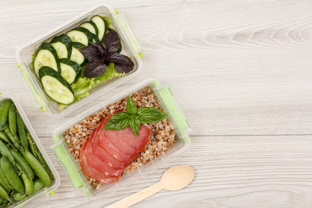 Contenitori di plastica per la preparazione dei pasti con piselli, porridge di grano saraceno bollito e fette di carne, cetrioli freschi e insalata, cucchiaio di legno su sfondo grigio. Vista dall'alto con copia spazio