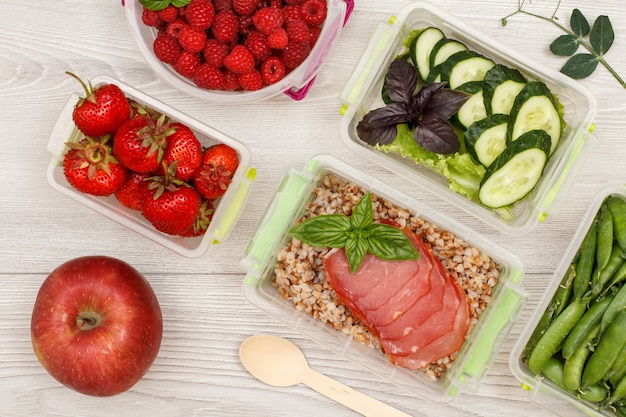 Contenitori per la preparazione di pasti in plastica e mele con fragole fresche, lamponi, porridge di grano saraceno bollito e fette di carne, cetrioli e insalata, piselli, cucchiaio di legno su sfondo grigio. Vista dall'alto