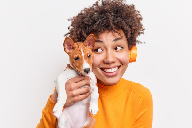 Content pleased African American girl smiles gently holds small puppy closely to face while walking in park happy to spend free time with pet expresses positive emotions isolated over white wall