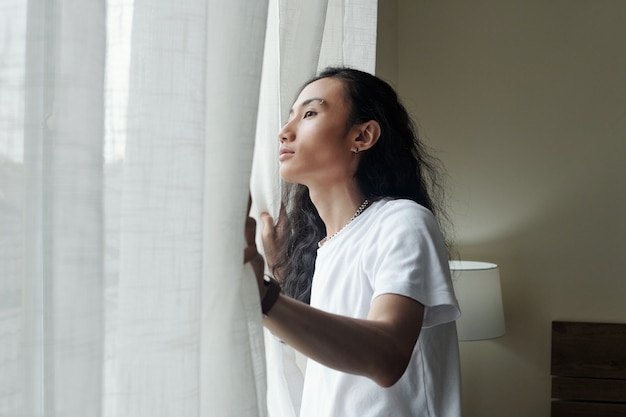 Contento giovane vietnamita con i capelli ondulati che aprono le tende mentre guarda fuori dalla finestra in hotel