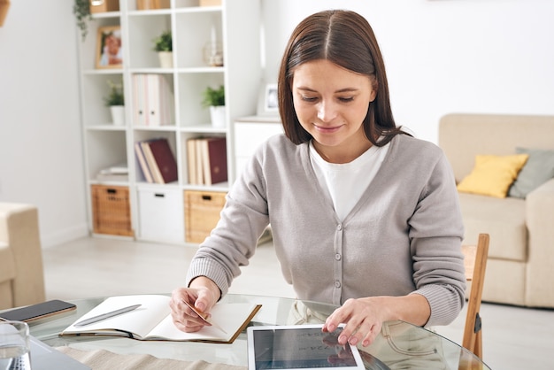 Contenuto attraente giovane donna in cardigan seduto al tavolo in soggiorno e utilizzando tablet app durante la gestione di denaro online