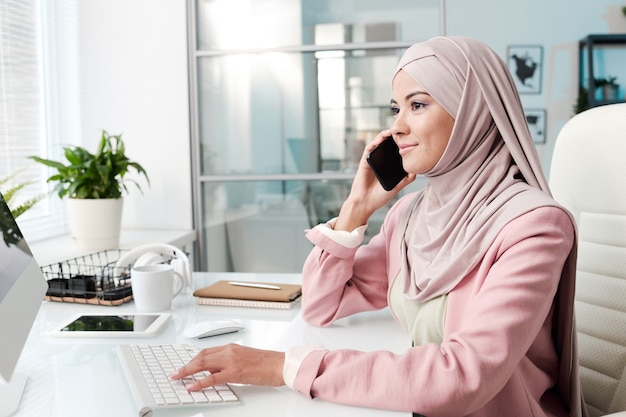 Contenuto attraente giovane imprenditrice musulmana in bellissimo hijab che comunica tramite telefono cellulare in ufficio