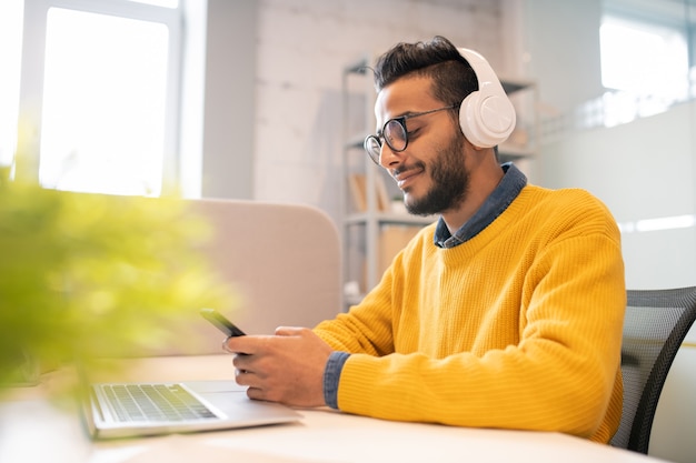 Contenuto soddisfatto giovane arabo con la barba seduto in ufficio moderno e messaggistica in smartphone mentre si lavora in ufficio