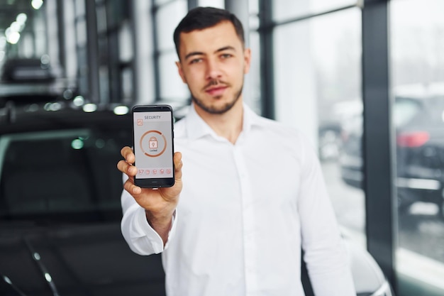 Contiene il telefono con etichette e icone Giovane uomo in camicia bianca è al chiuso con una nuova automobile moderna Telecomando