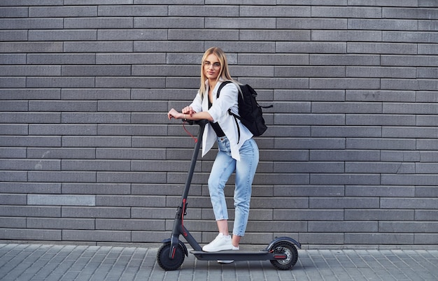 Contro il muro grigio Bella bionda in abiti casual che guidano uno scooter elettrico all'aperto durante il giorno soleggiato