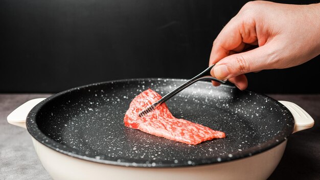 Controfiletto di manzo Wagyu A5 Premium Rare Slices con struttura ad alta marmorizzazione raccogliere e grigliare su una padella antiaderente rivestita di marmo con una pinza per barbecue con la mano su sfondo nero. Servito per Yakiniku.