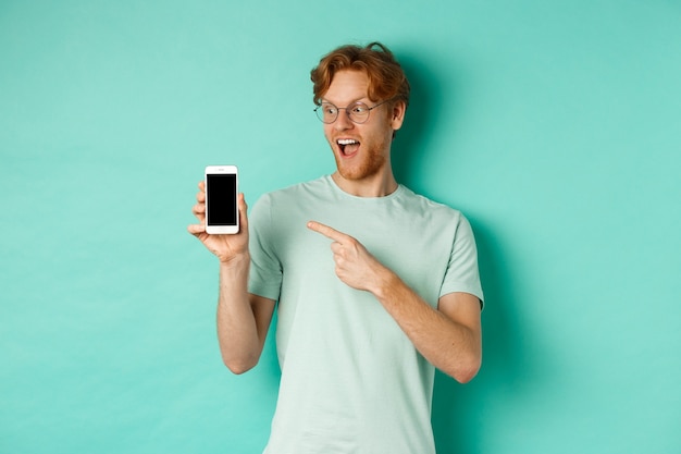 Controllalo. Bel ragazzo dai capelli rossi con gli occhiali che punta il dito sullo schermo vuoto dello smartphone, mostrando la promozione online, in piedi stupito su sfondo turchese