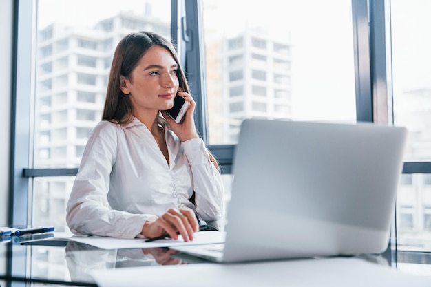 Conversare al telefono Giovane donna in abiti formali bianchi è al chiuso nell'ufficio moderno