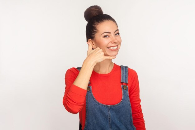 Conversazione telefonica Ritratto di ragazza carina ed elegante positiva con crocchia per capelli in tuta di jeans che mostra chiamami gesto con la mano a forma di telefono studio indoor girato isolato su sfondo bianco