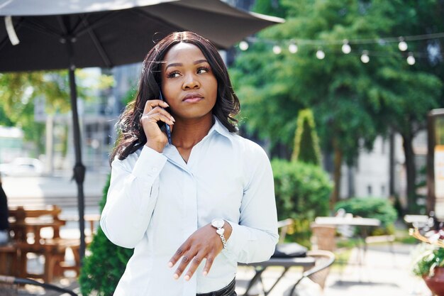 Conversazione tramite telefono Giovane donna afro americana in camicia bianca all'aperto in città vicino a alberi verdi e contro la costruzione di affari