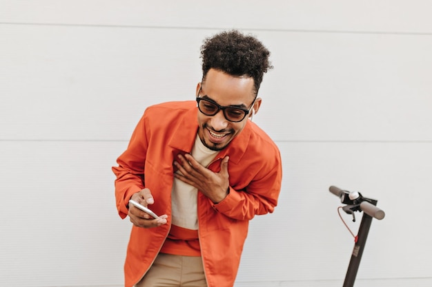 Cool brunet ricci uomo dalla pelle scura in giacca arancione occhiali da sole e pantaloncini beige ride tiene il telefono e posa vicino allo scooter fuori