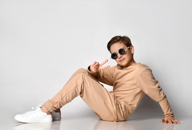 Cool preteen boy bambino in abbigliamento sportivo casual e occhiali da sole alla moda che si sente felice mostrando il gesto di vsign seduto sullo sfondo della parete dello studio e guardando la fotocamera Elegante ragazzo maschio bello positivo