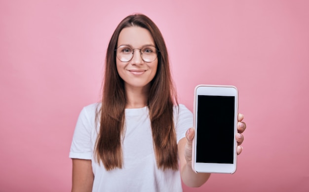 Cool ragazza allegra in una maglietta bianca in piedi e mostra il display del telefono proprio nel telaio