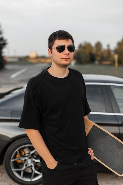 Cool uomo modello hipster in maglietta nera alla moda con longboard si trova vicino a un'auto all'aperto