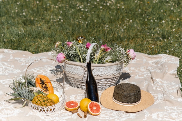 Coperta da picnic su un prato con cesto pieno di fiori, frutti tropicali, un cappello di paglia e una bottiglia di vino.