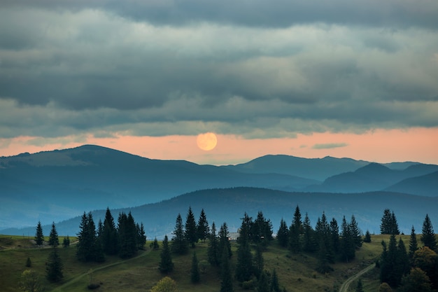 Coperto di montagne nebbiose al sorgere della luna, grande luna sul cielo arancione brillante sopra alti abeti.