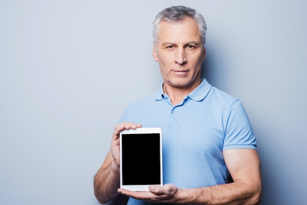 Copia spazio sul suo tablet. Fiducioso uomo anziano che mostra la sua tavoletta digitale e sorride mentre si trova in piedi su sfondo grigio