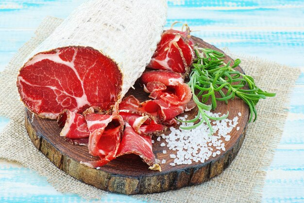 Coppa stagionata affettata con spezie e un rametto di rosmarino su fondo rustico in legno.