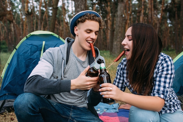 Coppia affamata mangia salsicce e beve birra vicino al falò. Romanticismo turistico. Stile di vita da campeggio.
