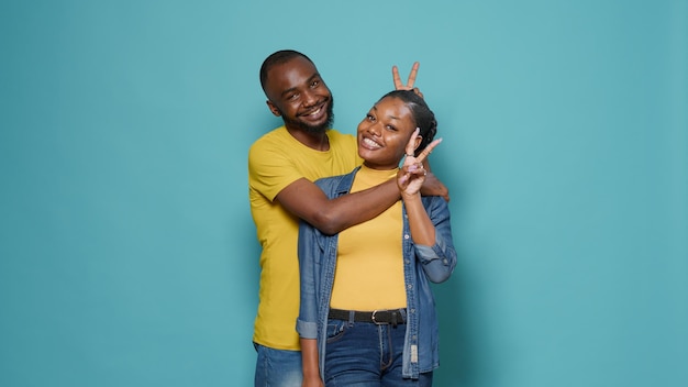 Coppia afroamericana in un abbraccio che mostra il segno di pace con le mani, in piedi su sfondo blu. Uomo e donna che scherzano e fanno pubblicità a un gesto pacifico insieme in studio.