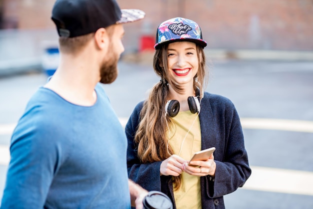 Coppia alla moda in maglioni blu e berretti che parlano insieme in piedi con lo skateboard all'aperto nel parco giochi sul tetto