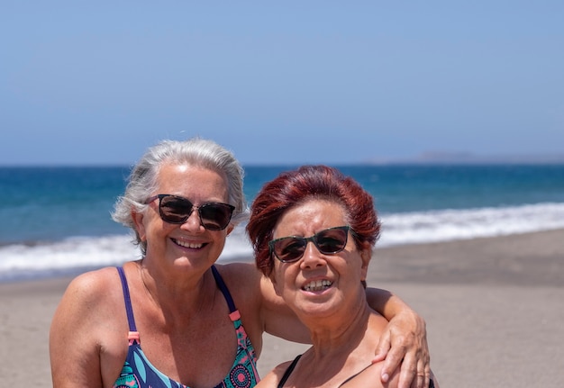 Coppia allegra di donne anziane in amicizia che si godono la spiaggia insieme sotto il sole con onde e mare blu sullo sfondo - pensionati attivi e concetto di vacanza