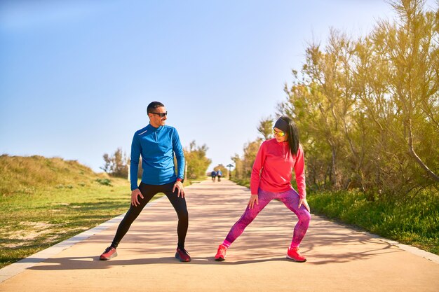 Coppia allungando le gambe prima dell'allenamento. La donna indossa abiti rosa brillante e viola. L'uomo indossa una camicia blu e pantaloni lunghi neri.