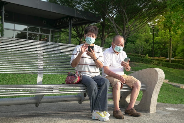 Coppia asiatica anziana che indossa una maschera facciale seduta e legge i loro telefoni in un parco
