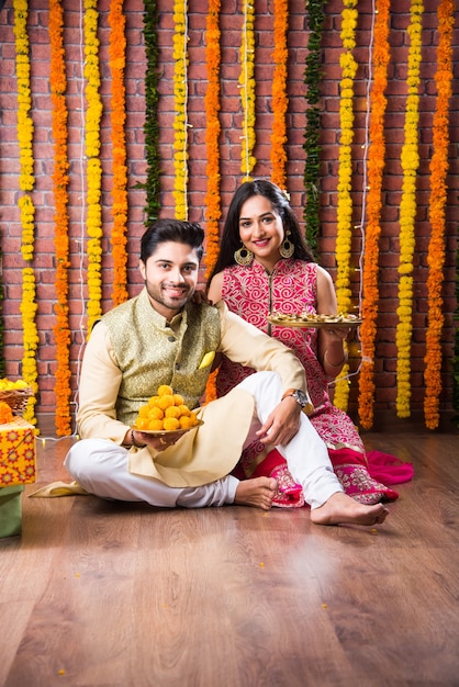 Coppia attraente indiana in abbigliamento tradizionale che celebra il Diwali Festival, il compleanno o l'anniversario con regali a sorpresa e dolce laddoo su sfondo decorato con fiori di calendula