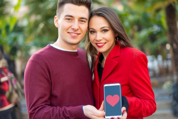 Coppia baciare mentre mostrano uno smartphone con una forma del cuore, datando il concetto dell'applicazione