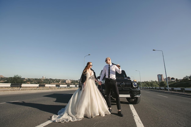 Coppia brutale la sposa e lo sposo con un pipistrello in giacche di pelle vicino all'auto sulla strada