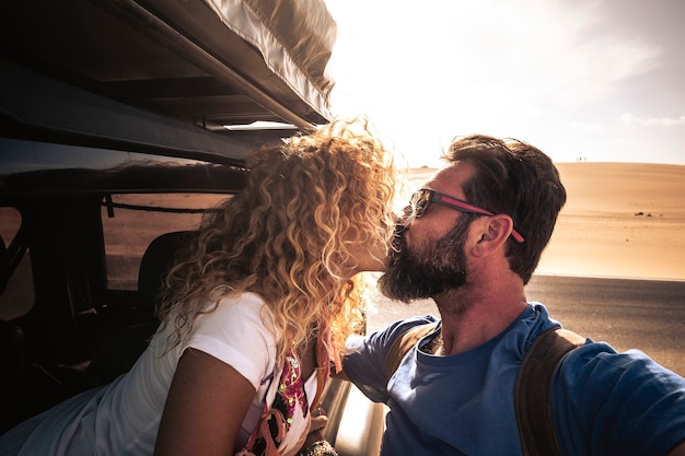 Coppia carina caucasica adulta di viaggiatori si baciano con il sole in controluce - auto che viaggia insieme all'amore e alla relazione - deserto sabbioso in backgrounf e cielo luminoso - uomo con la barba
