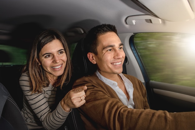 Coppia caucasica all'interno dell'auto sorridente. La donna sta indicando qualcosa fuori con il dito mentre l'uomo sta guidando.