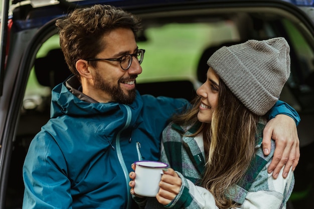 Coppia caucasica millenaria soddisfatta seduta nel bagagliaio dell'auto nella foresta fredda goditi l'avventura e una tazza di bevanda calda