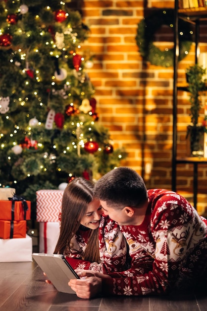 Coppia caucasica uomo e donna che riposa a casa vicino all'albero di capodanno incandescente e digitando sul tablet