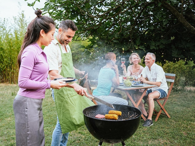 Coppia che cucina su un barbecue in campagna Sul retro i suoi amici festeggiano con bevande alcoliche