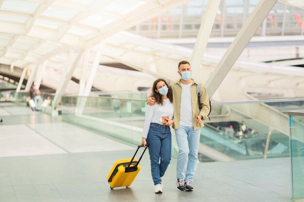 Coppia con maschere mediche che cammina attraverso il moderno terminal dell'aeroporto con i bagagli